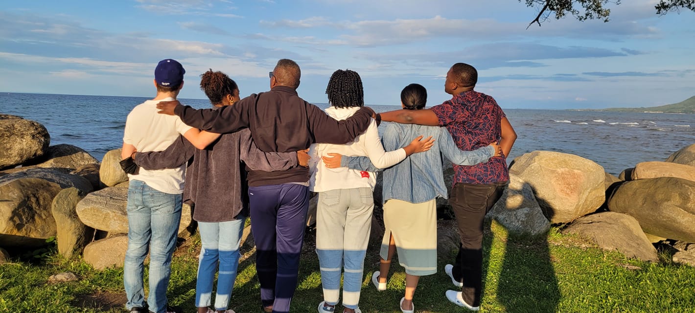 Six family members stand arm in arm on a rocky shoreline, looking out over the water together.