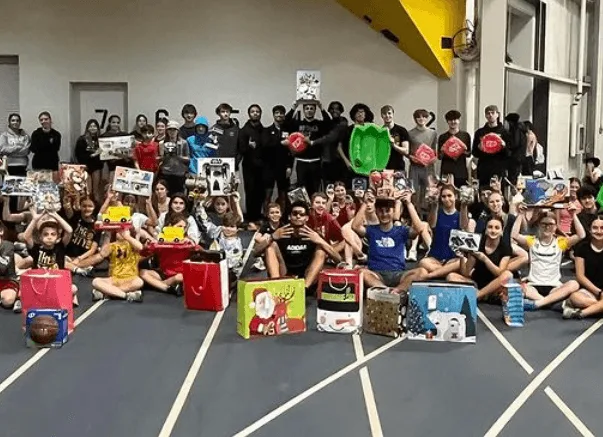 Students seated in a gymnasium behind rows of wrapped gifts and donation bags collected for the holiday drive.