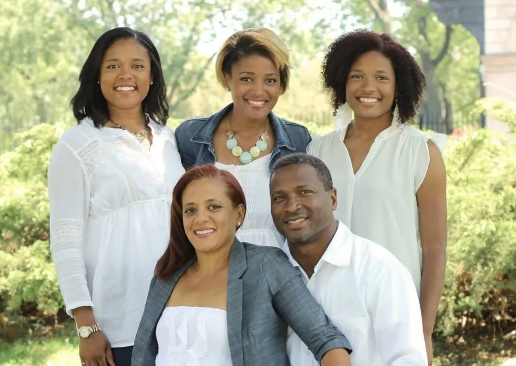 A family of five dressed in white and denim, gathered outdoors among summer greenery.
