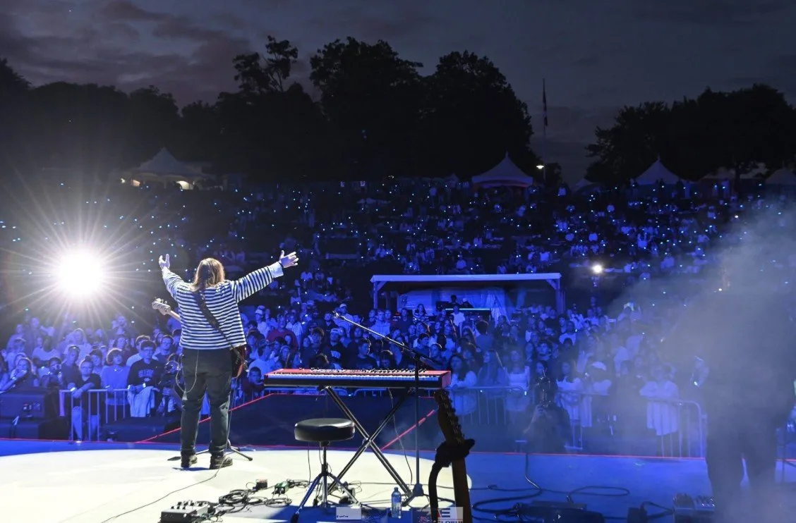 Leeland performing on an outdoor stage at dusk, arms raised in front of a large crowd lit by stage lights.
