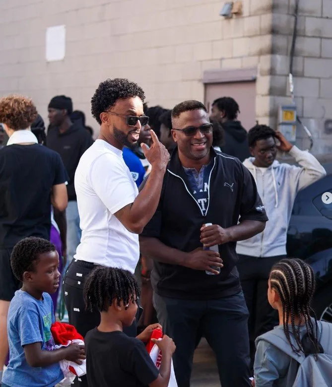 Two men laughing outdoors at the MH100 backpack giveaway while children in the foreground carry gift bags.