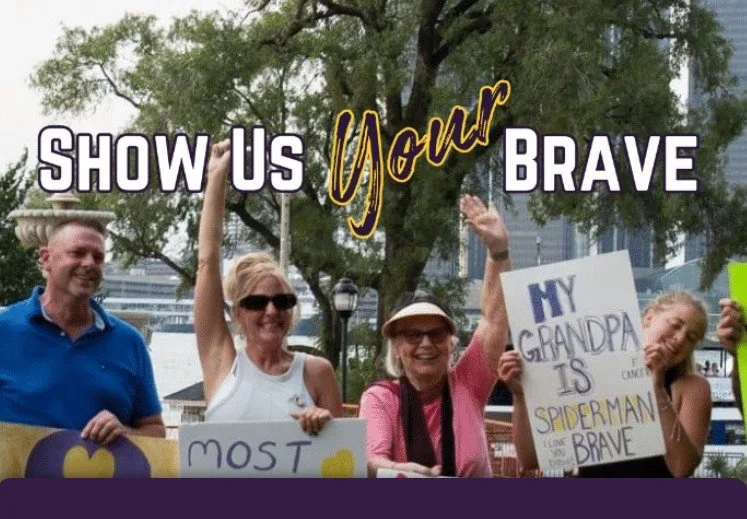 Supporters outdoors holding handmade signs under the words Show Us Your Brave, one sign reading My grandpa is Spiderman brave.