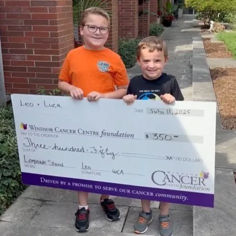 Two young boys on a driveway holding an oversized cheque for $350 made out to the Windsor Cancer Centre Foundation, with lemonade stand written in the memo line.