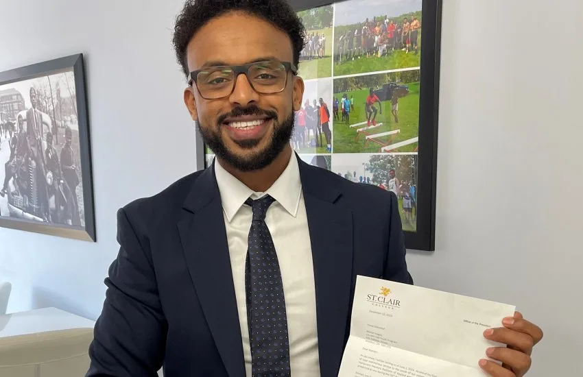 Mehari Hagos in a suit holding his letter from St. Clair College, with a collage of MH100 programme photographs behind him.
